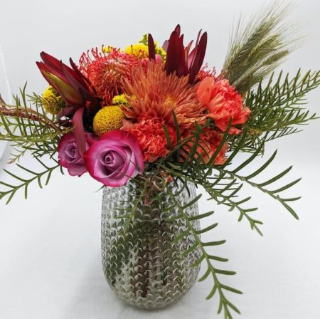 The bold colors of fall come together in this bouquet. Orange pincushion protea, lavender roses, bronze mums, orange carnations, wheat, yellow craspedia and lots of textural elements and greenery are designed in a hand-tied fashion and presented in a beautiful pebble textured glass vase. A perfect floral gift for fall birthdays and parties. We do our best to replicate our designs, please understand reasonable substitutions may be made.